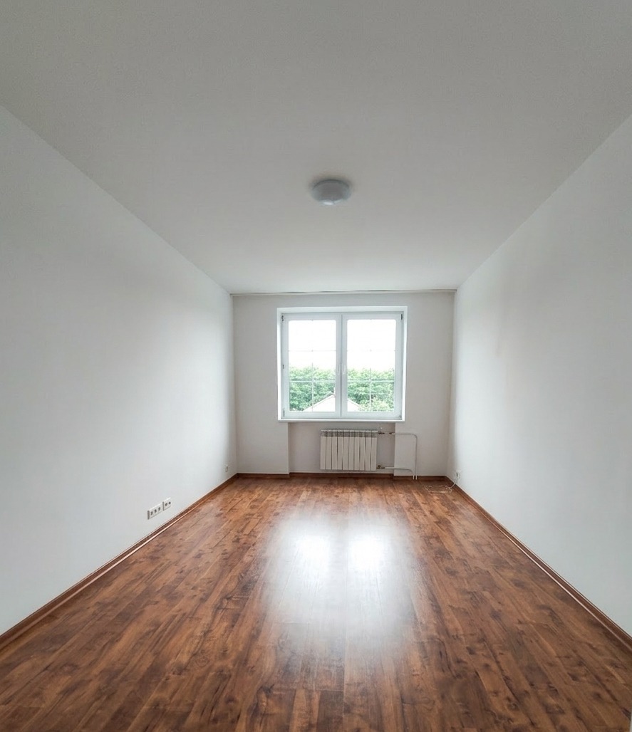 Puste, odświeżone wnętrze pokoju z drewnianą podłogą, białymi ścianami i oknem z widokiem na zieleń. Grzejnik pod oknem. Minimalistyczne wykończenie.