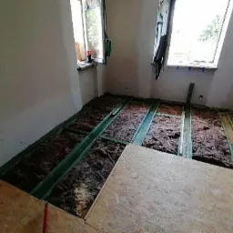 Poddasze w trakcie izolacji: widoczne zielone legary, brązowa wełna mineralna pomiędzy nimi oraz częściowo ułożone płyty OSB. Dwa okna dachowe w tle. Prace w toku.