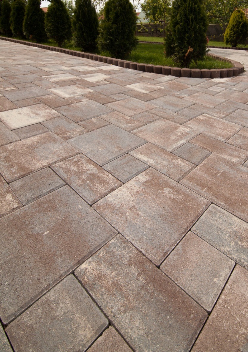 Ułożona nawierzchnia z kostki brukowej w odcieniach szarości i brązu, obrzeże z małych betonowych elementów, rząd tuj i trawnik w tle.