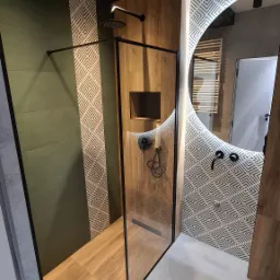 Nowoczesna łazienka z kabiną prysznicową typu walk-in, wykończona płytkami w odcieniach zieleni, drewna i bieli z geometrycznym wzorem; widoczna armatura w kolorze czarnym.