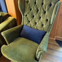 Górska Opieka Nieruchomości Paweł Strzępek - Zielony, welurowy fotel typu uszak z pikowanym oparciem i granatową poduszką, stojący na drewnianej podłodze. Widoczny fragment drewnianej boazerii.