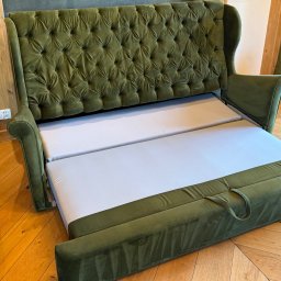 Górska Opieka Nieruchomości Paweł Strzępek - Rozłożona sofa w odcieniach zieleni z pikowanym oparciem, ukazująca biały materac. Widoczna drewniana podłoga i fragment ściany w tle. Sofa rozłożona do spania.
