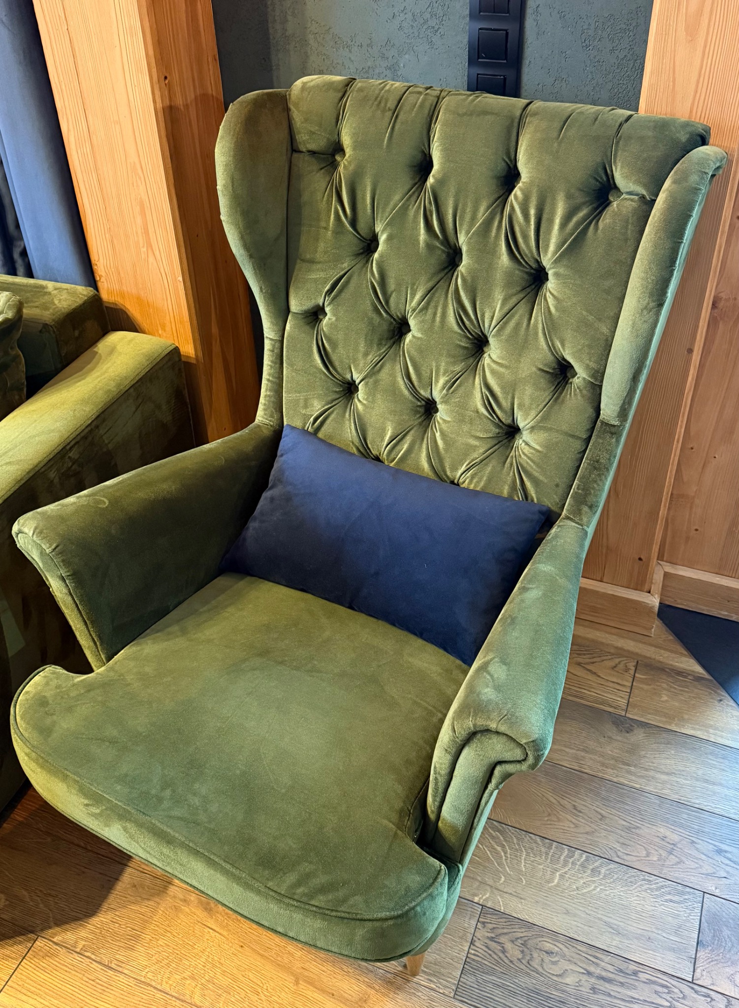 Zielony, welurowy fotel typu uszak z pikowanym oparciem i granatową poduszką, stojący na drewnianej podłodze. Widoczny fragment drewnianej boazerii.