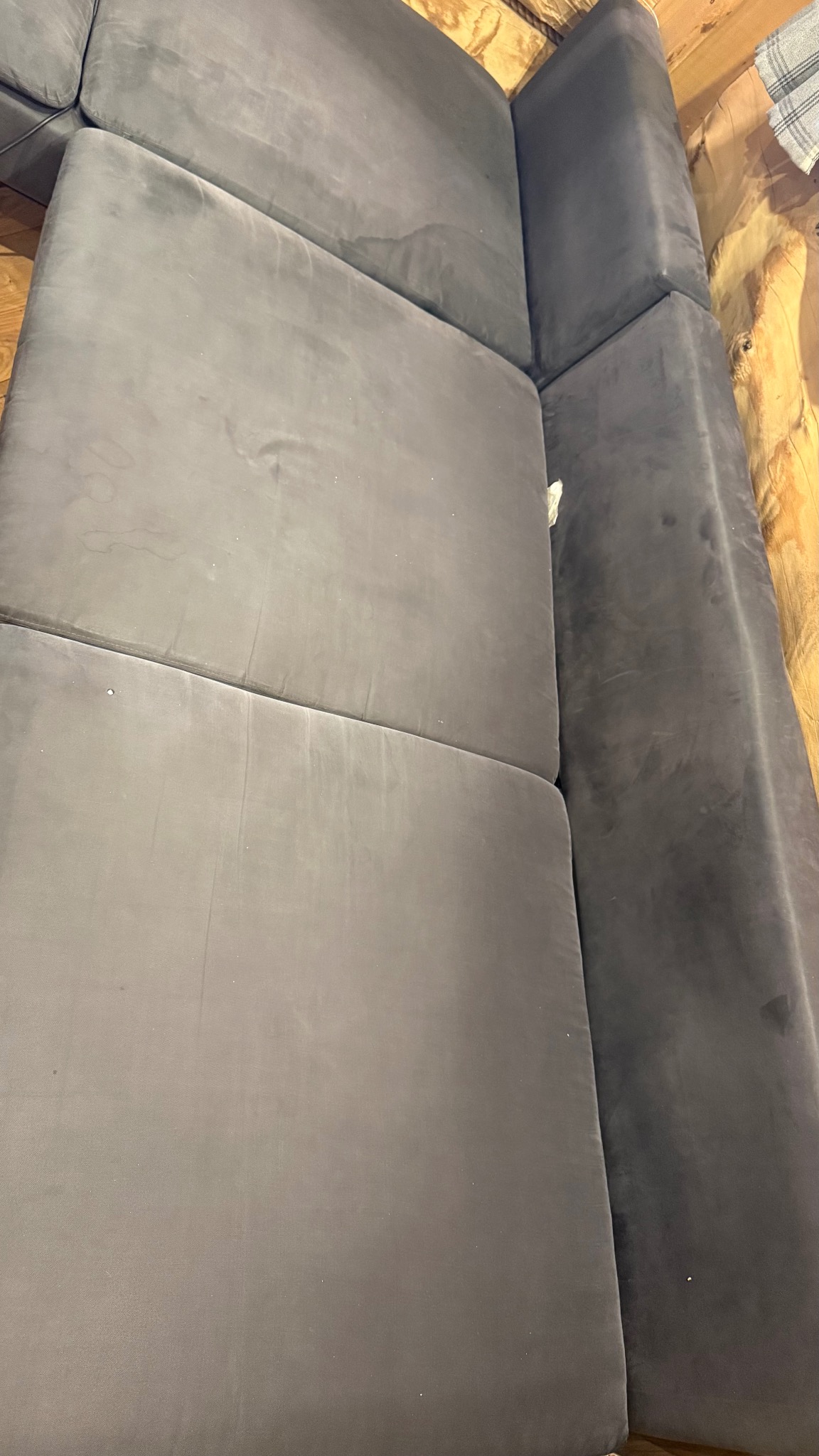 Szary, modułowy narożnik z welurową tapicerką, widoczny z góry, z delikatnymi śladami użytkowania. Tło stanowi drewniana ściana z widocznymi sękami.