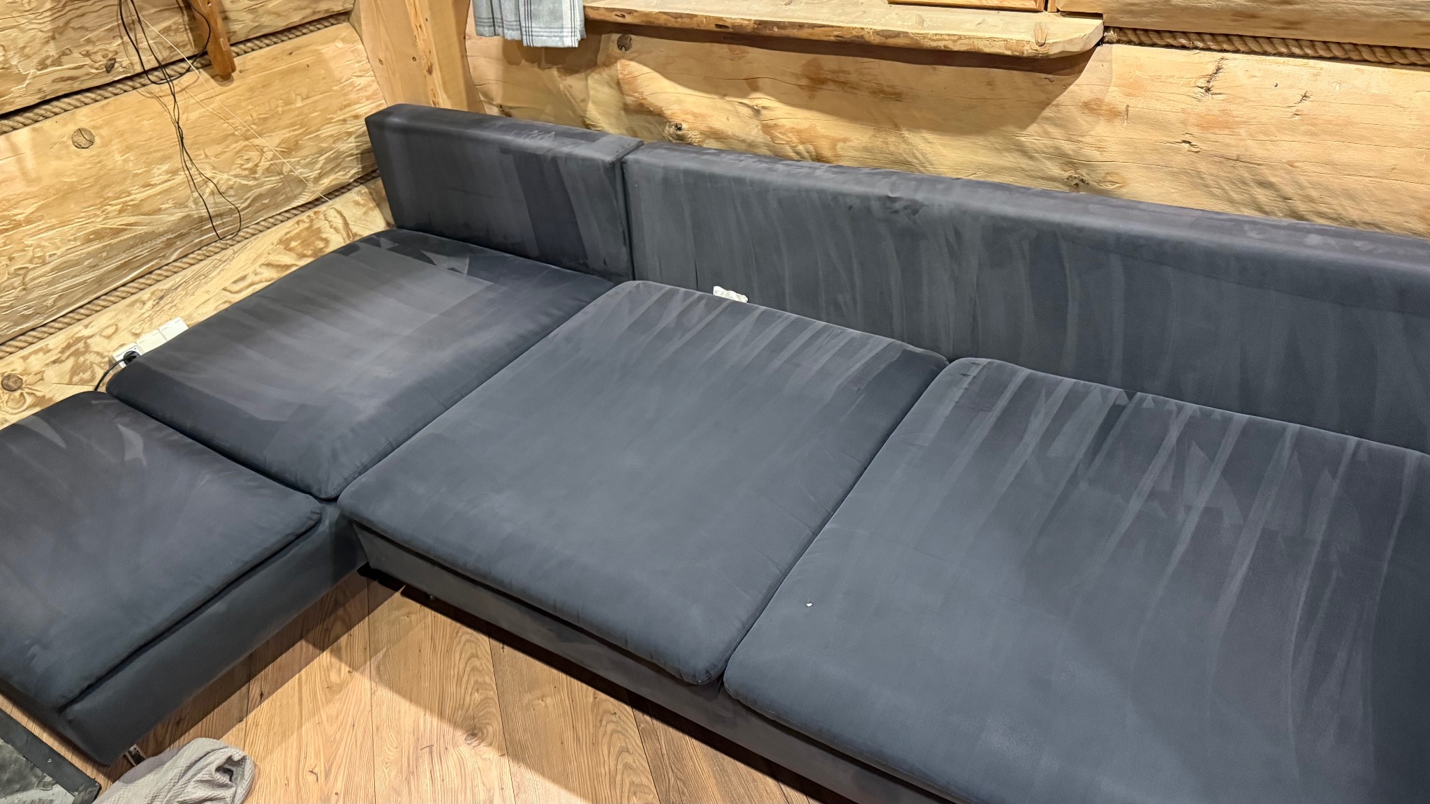 Szara, narożna kanapa po praniu, widoczne ślady po czyszczeniu na tle drewnianej ściany. Ujęcie z góry, ukazujące detale faktury materiału i świeżość po zabiegu.