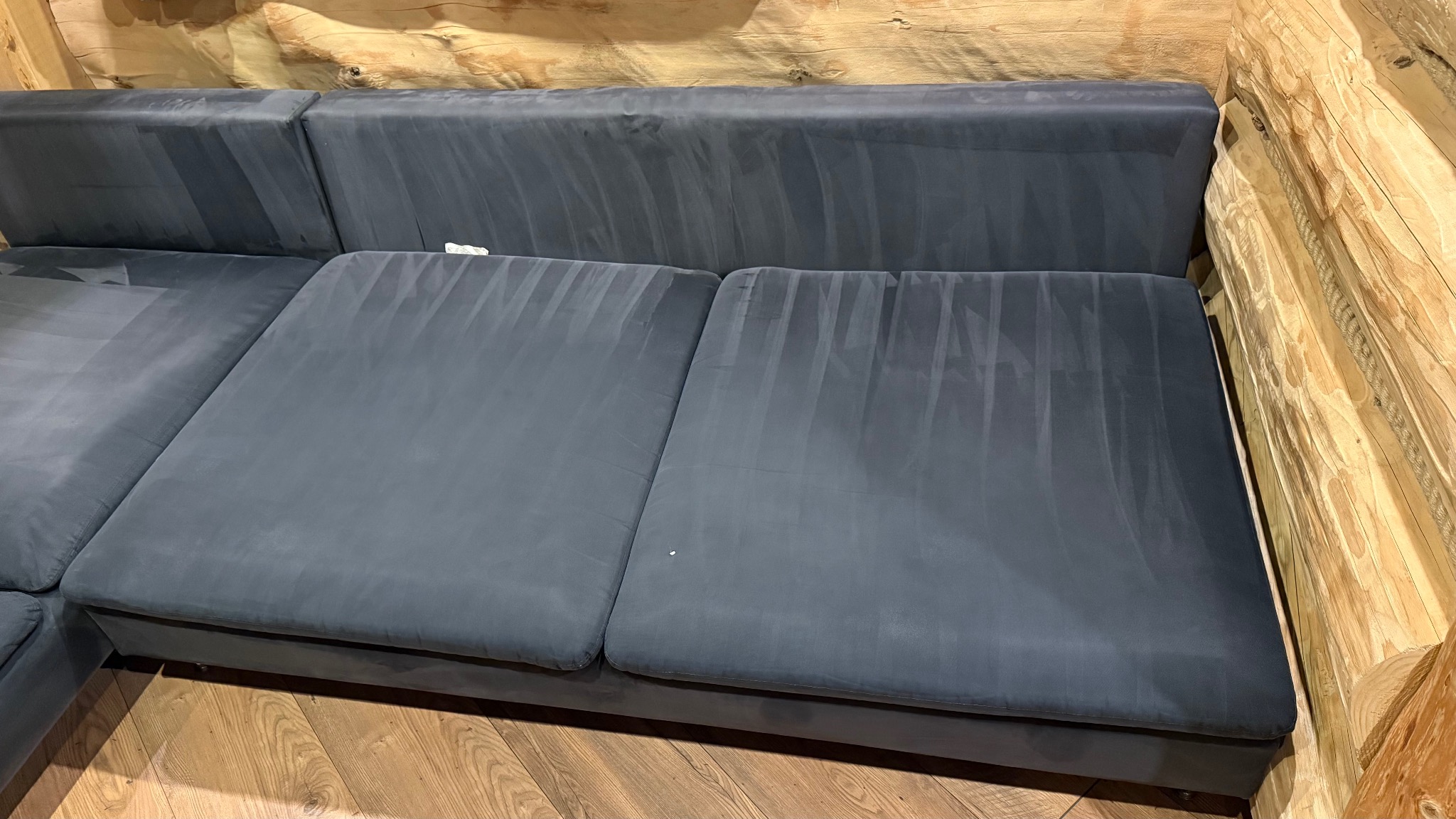 Szara, narożna kanapa po praniu ekstrakcyjnym, widoczne pionowe ślady po czyszczeniu. Tło: drewniana ściana z bali i podłoga z paneli.