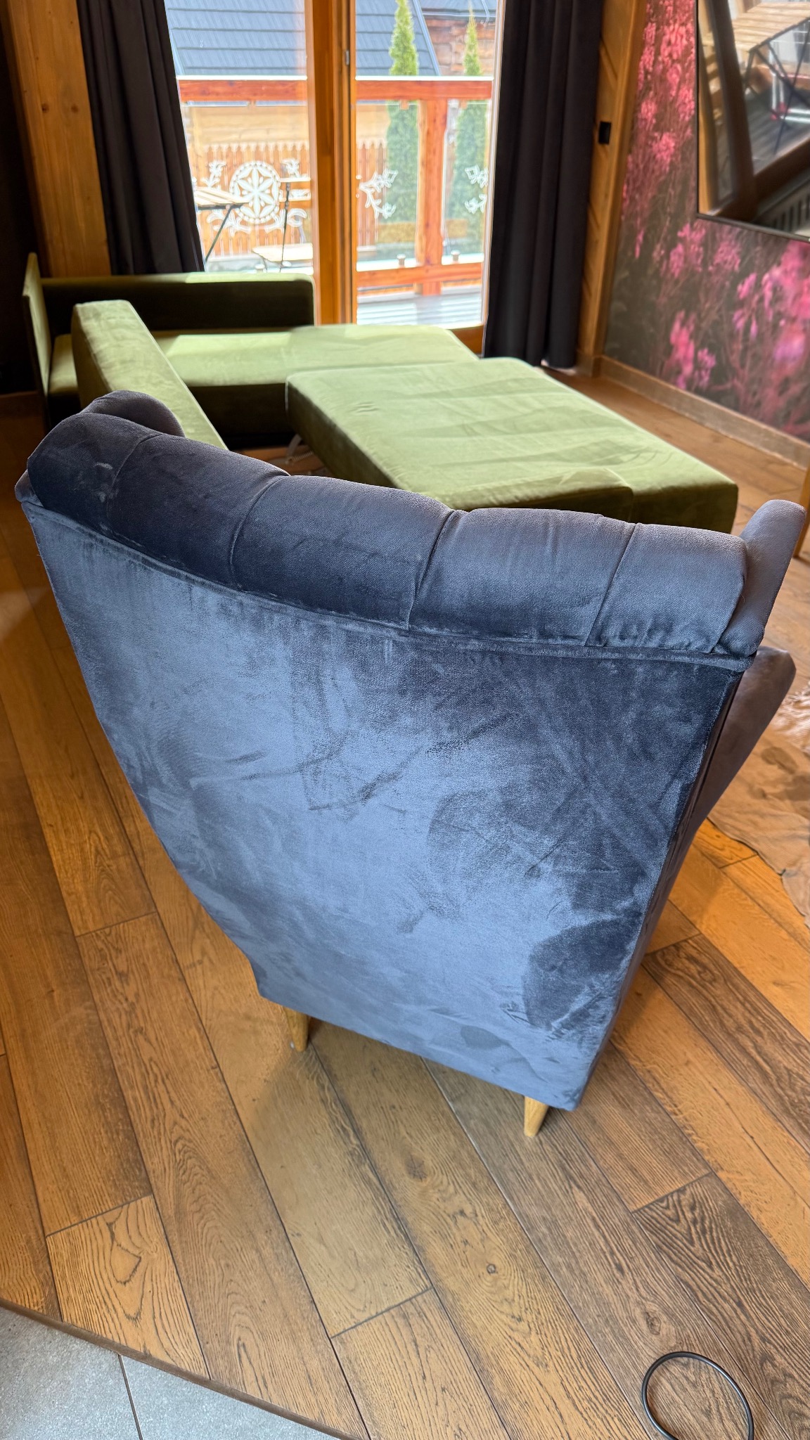 Szary, welurowy fotel na drewnianej podłodze, z rozłożoną zieloną sofą w tle. Widok z tyłu fotela, z oknem i dekoracyjną ścianą w tle.