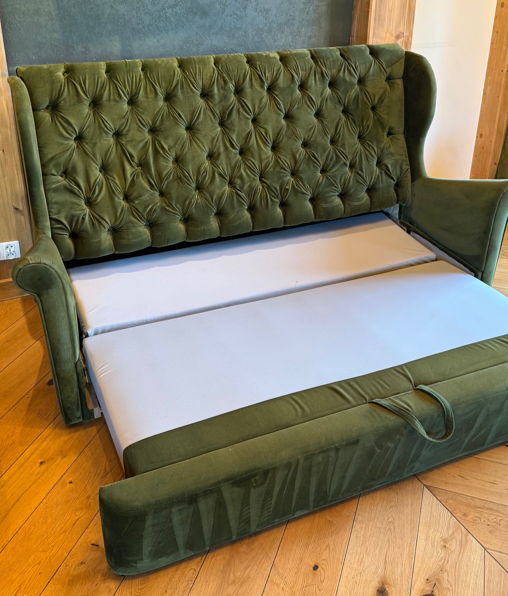 Rozłożona sofa w odcieniach zieleni z pikowanym oparciem, ukazująca biały materac. Widoczna drewniana podłoga i fragment ściany w tle. Sofa rozłożona do spania.
