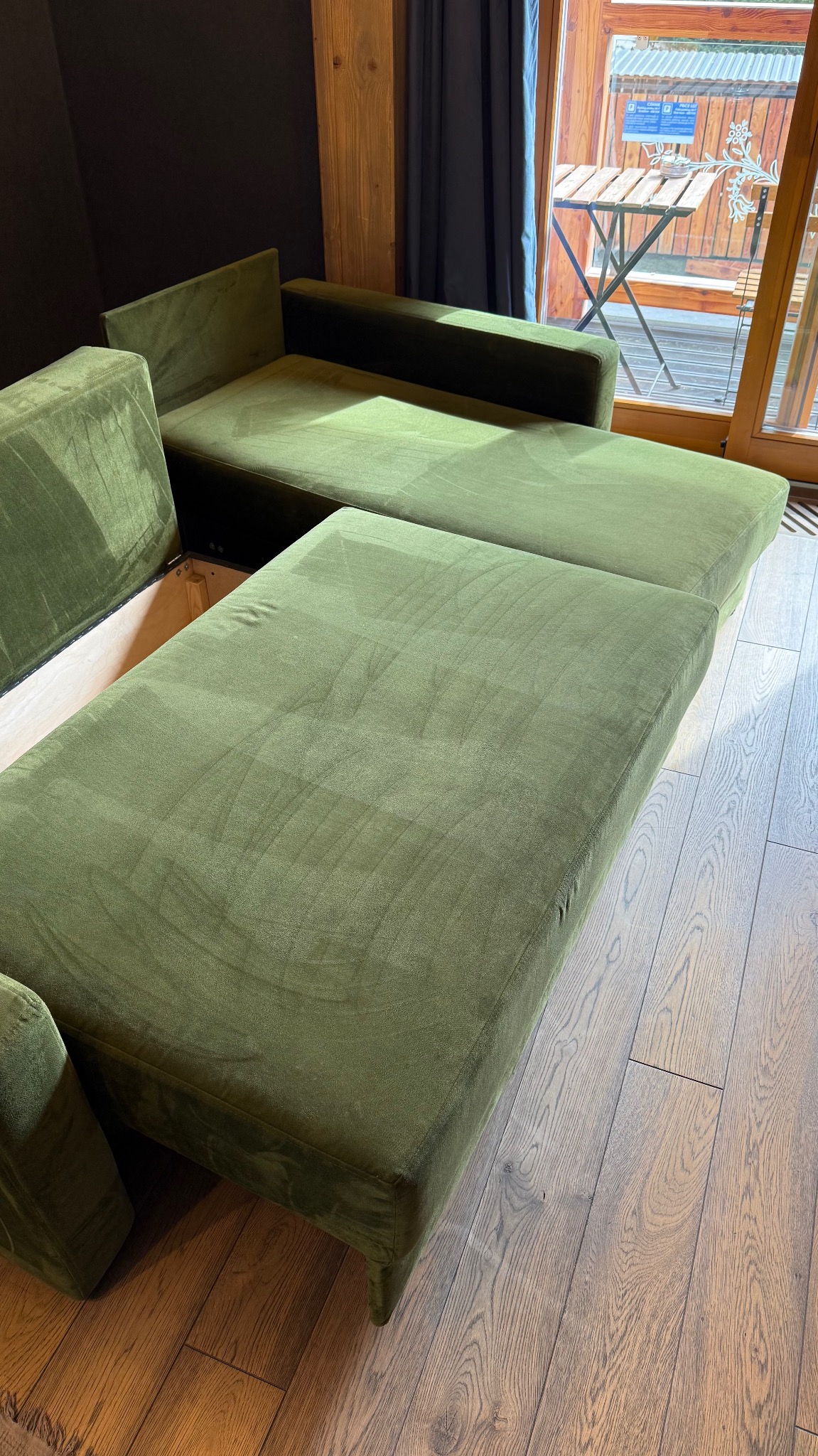 Rozłożysta sofa w odcieniach zieleni na drewnianej podłodze, z widokiem na balkon przez okno. Minimalistyczny design i naturalne materiały.