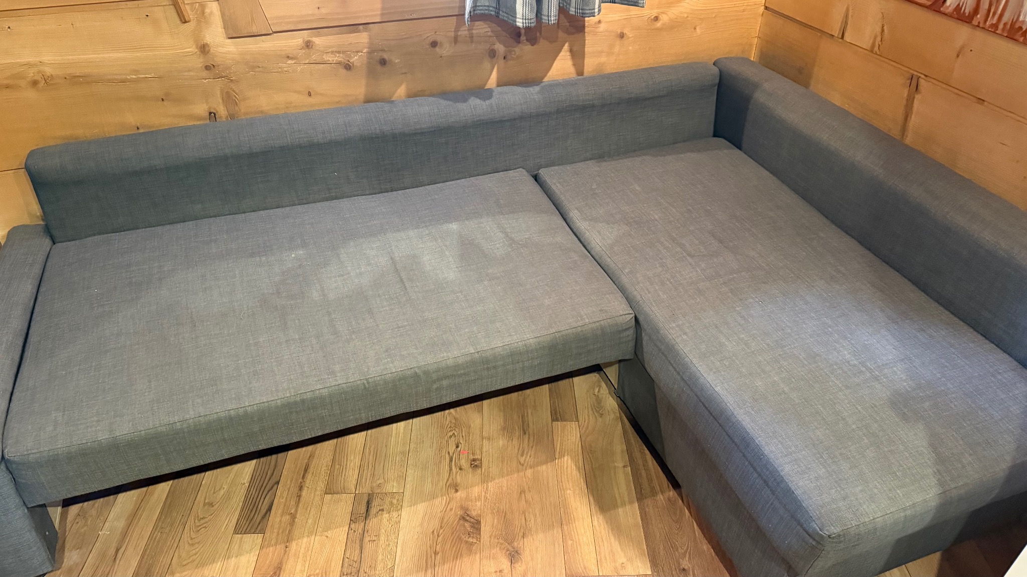 Szara, narożna kanapa z tkaniny w rustykalnym wnętrzu z drewnianą boazerią. Widok z góry, ukazujący fakturę materiału i kształt mebla.