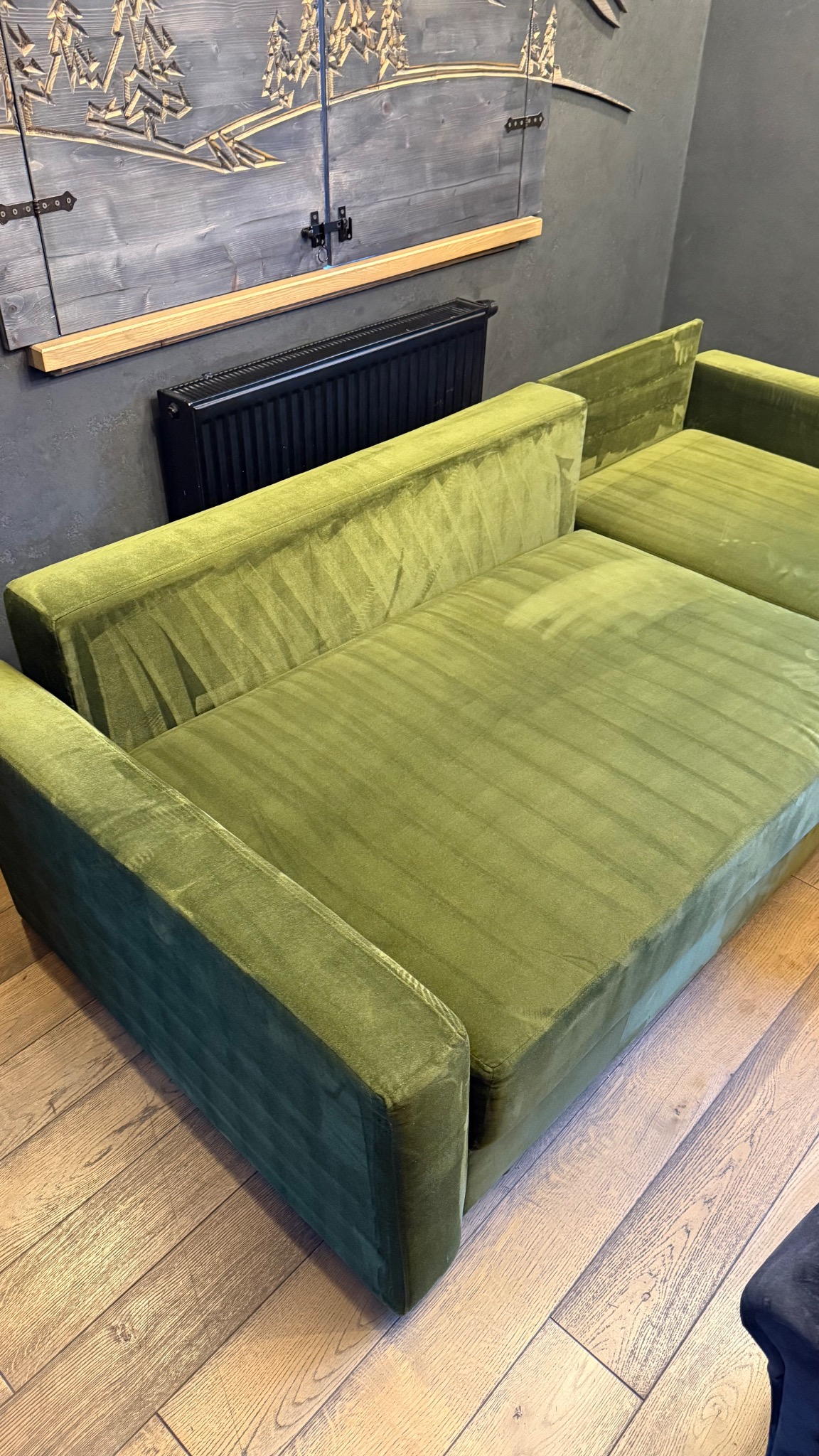 Sofa modułowa w odcieniach zieleni, obita welurem, na tle szarej ściany z dekoracyjną, drewnianą płaskorzeźbą i czarnym grzejnikiem.