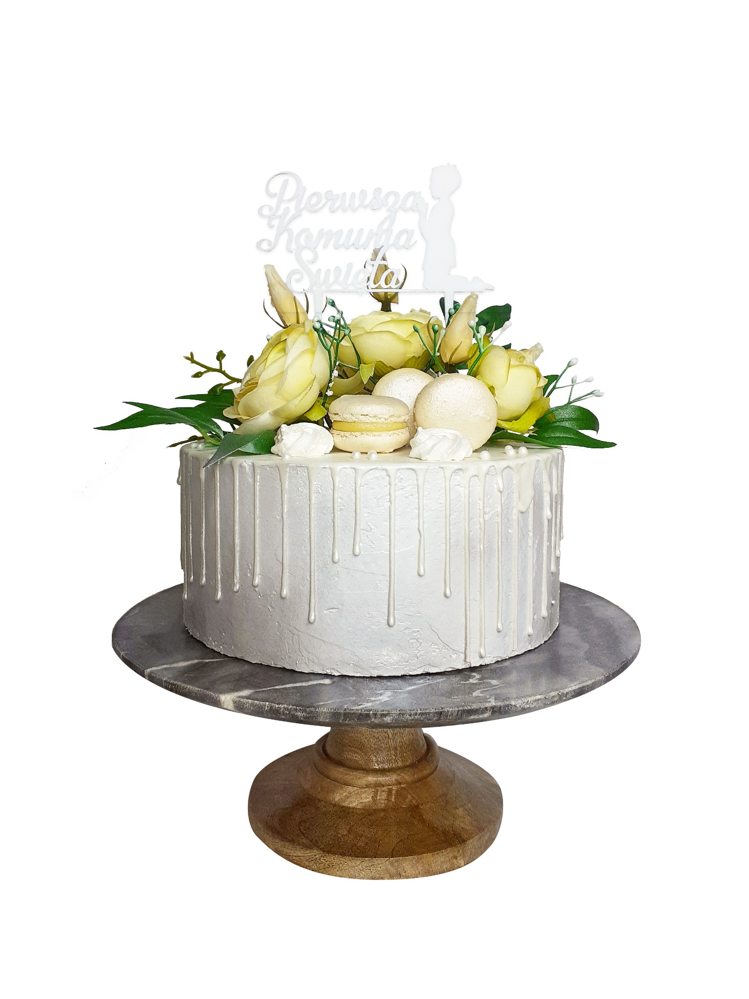 Tort komunijny z białym lukrem spływającym po bokach, dekorowany żółtymi różami, makaronikami i napisem 'Pierwsza Komunia Święta' na drewnianym stojaku.