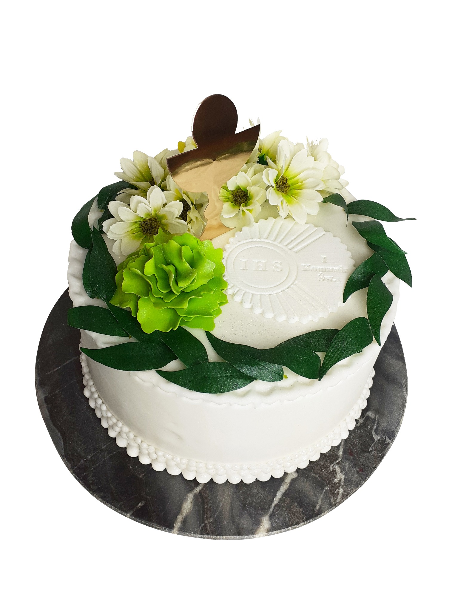 Tort komunijny z białym lukrem, dekorowany zielonymi liśćmi, białymi kwiatami i złotym krzyżem, umieszczony na marmurowej podstawie.