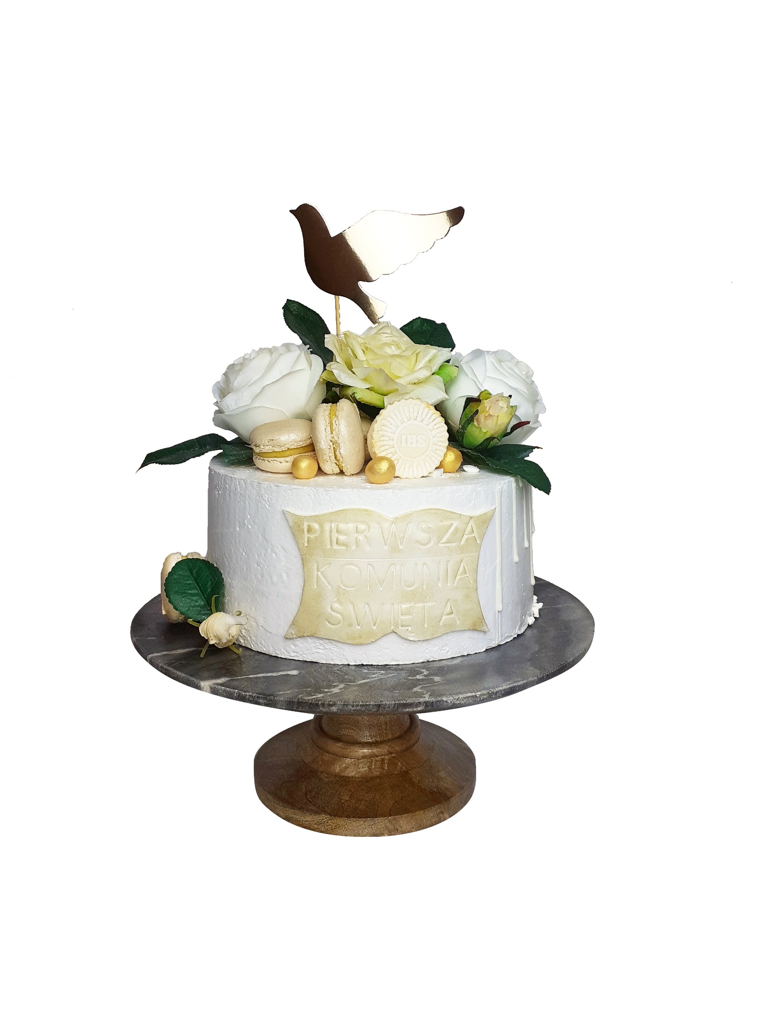 Tort komunijny z białym lukrem, dekorowany białymi i żółtymi różami, makaronikami, złotym gołębiem i napisem 'Pierwsza Komunia Święta', umieszczony na marmurowym stojaku.
