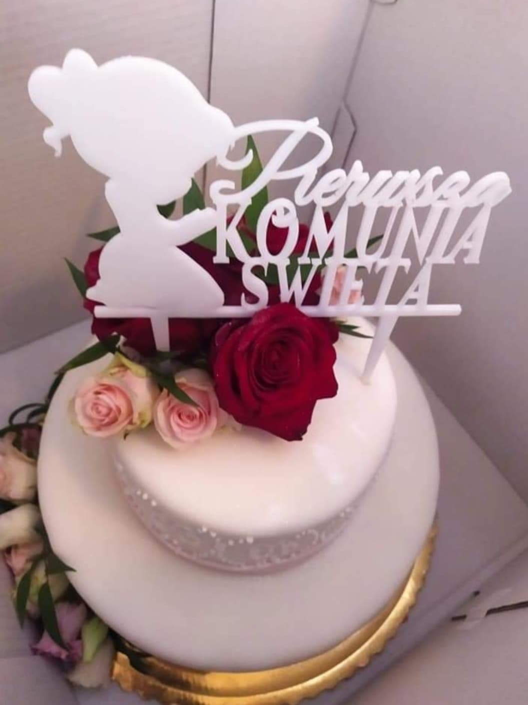 Dwupiętrowy tort z białym lukrem, dekorowany czerwonymi i różowymi różami oraz białym topperem z napisem 'Pierwsza Komunia Święta' i sylwetką dziewczynki w sukience.