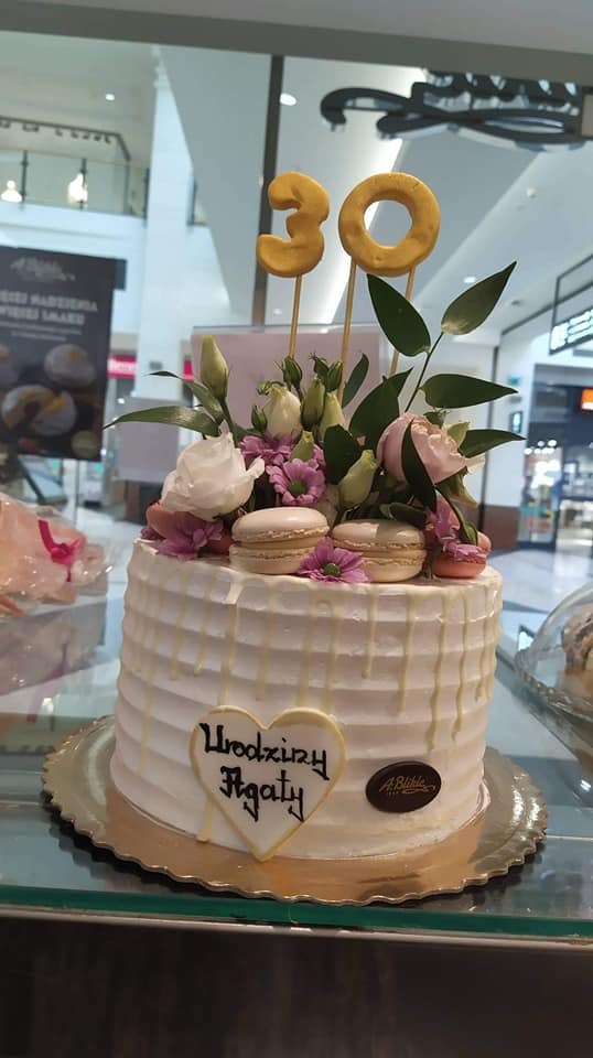 Tort urodzinowy z białym kremem, dekorowany kwiatami, makaronikami i złotymi cyframi 30, z napisem 'Urodziny Agaty' w kształcie serca, na złotej tacy, w tle wnętrze galerii handlowej.