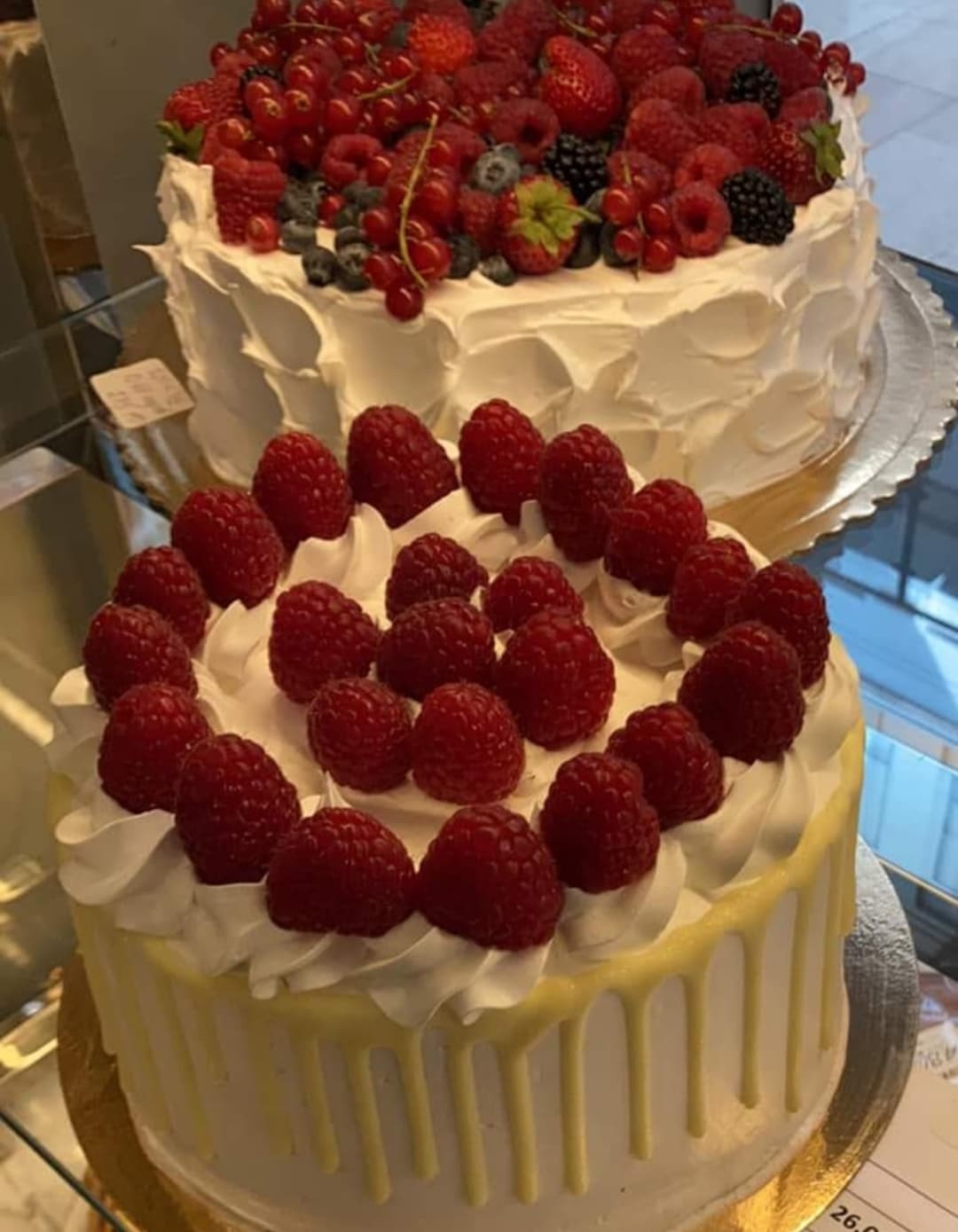 Dwa torty: jeden z malinami i białymi kremowymi rozetkami, drugi z mieszanką czerwonych porzeczek, malin, jeżyn i borówek, oba na srebrnych podkładkach.