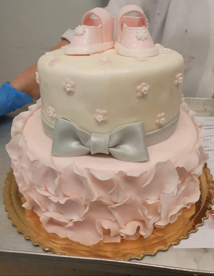 Dwupiętrowy tort w pastelowych kolorach, ozdobiony falbankami, srebrną kokardą i lukrowanymi bucikami niemowlęcymi na wierzchu.