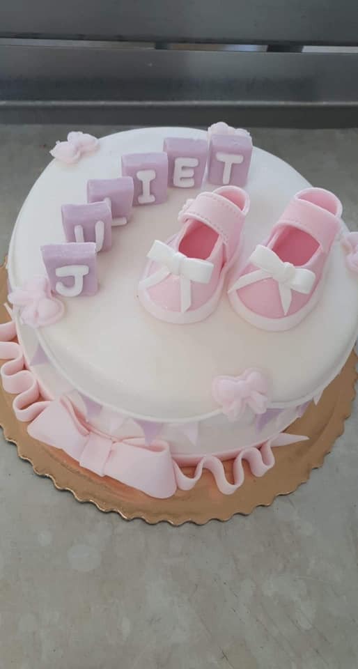 Tort dwupoziomowy z dekoracjami z masy cukrowej: różowe buciki niemowlęce z kokardkami i kostki z literami układającymi się w imię 'Juliet'.