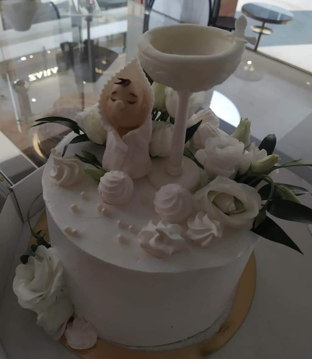 Tort na chrzest z białą polewą, dekorowany figurką niemowlęcia w beciku, białymi różami, bezami i kielichem z czekolady.