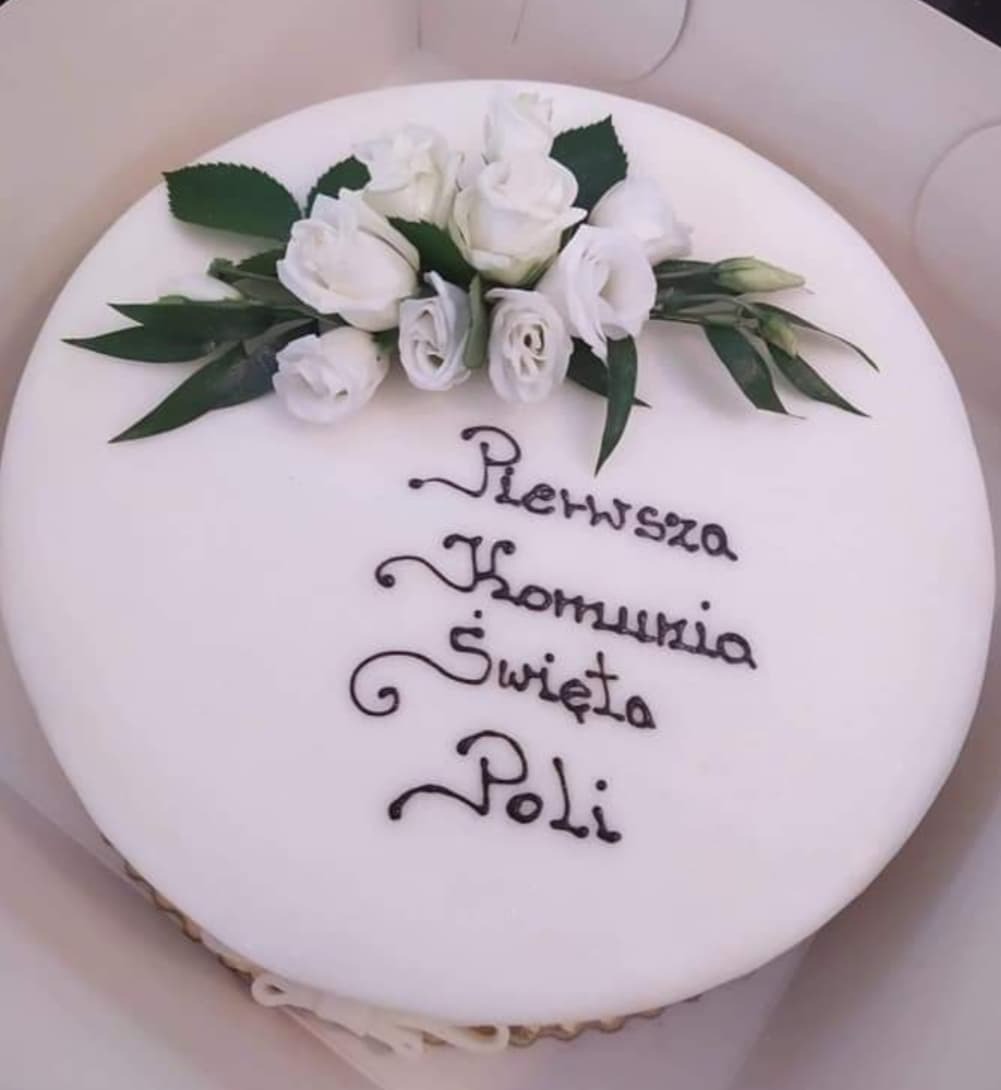 Okrągły tort z białym lukrem, udekorowany bukietem białych róż i zielonych liści, z napisem 'Pierwsza Komunia Święta Poli' wykonanym czarnym lukrem.