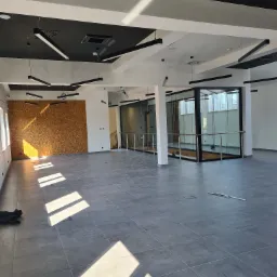 Przestronne wnętrze biurowe z szarymi płytkami podłogowymi, drewnianą ścianą akcentującą i nowoczesnym oświetleniem sufitowym. Widoczne fragmenty szklanych ścianek działowych i wpadające promienie...