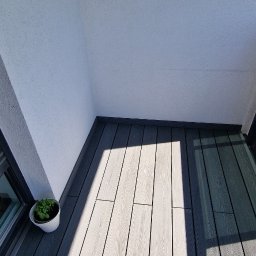 REMONTOZNAWCY - Nowoczesny balkon z szarym, kompozytowym deskowaniem. W lewym dolnym rogu doniczka z zieloną rośliną. Widoczne cienie tworzą geometryczne wzory na podłodze.