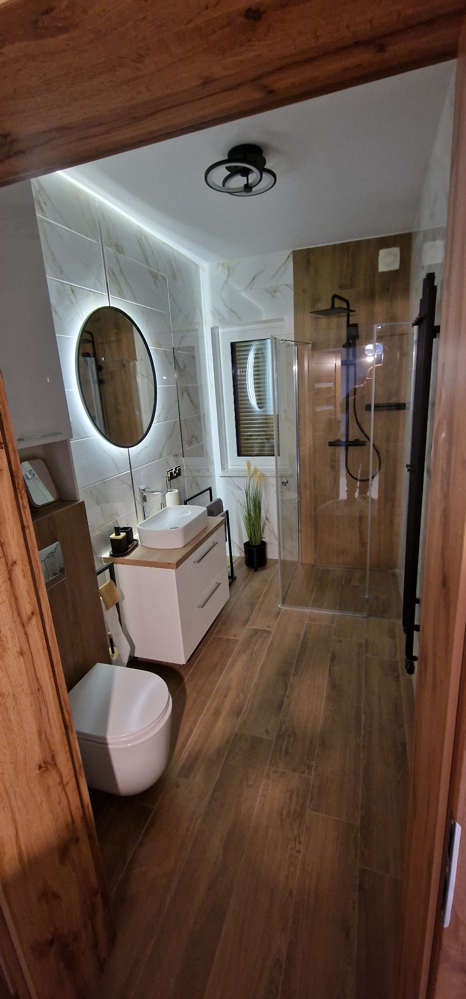 Nowoczesna łazienka z drewnopodobną podłogą, białą umywalką nablatową, okrągłym lustrem z podświetleniem LED i kabiną prysznicową typu walk-in z czarną armaturą.
