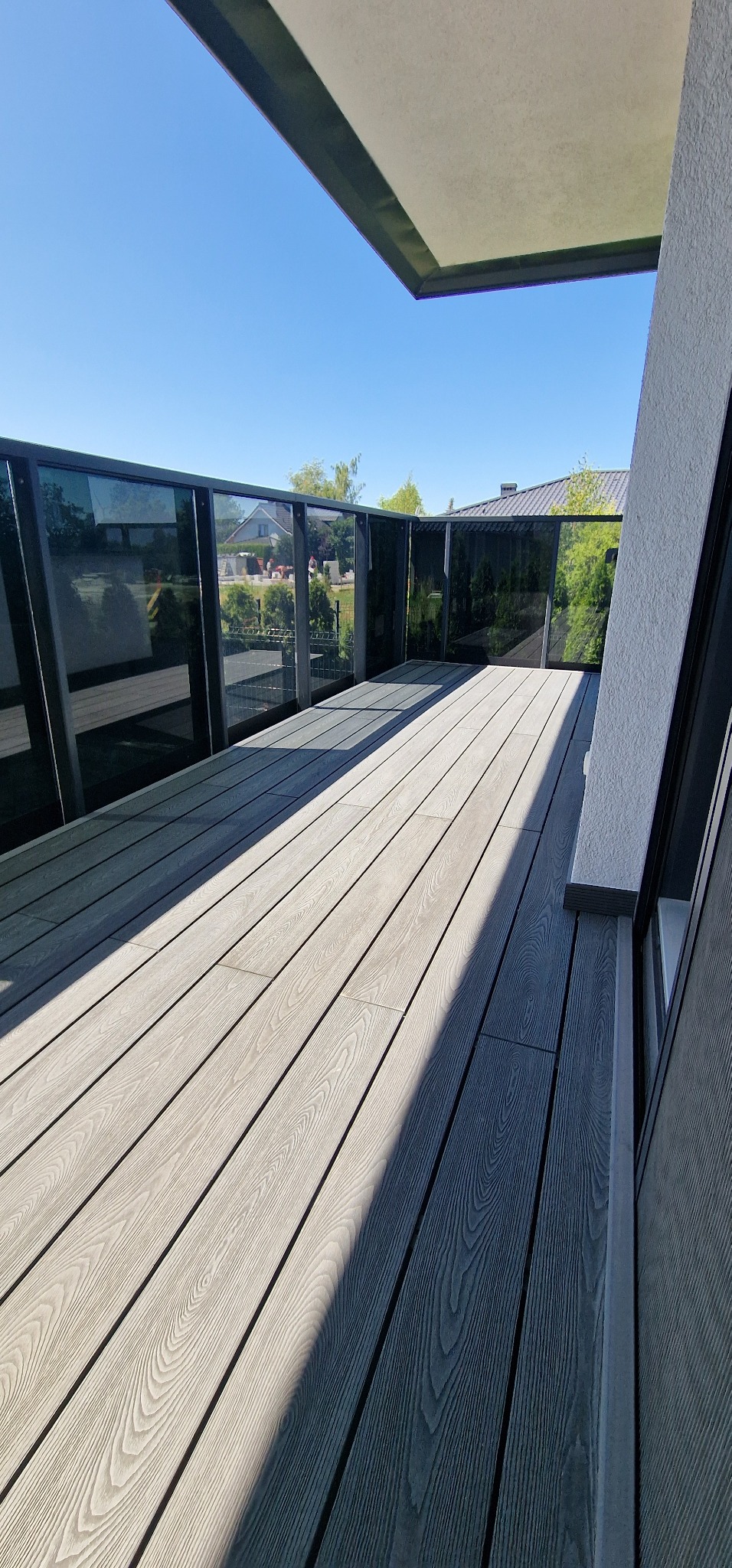 Nowoczesny balkon z szarymi deskami kompozytowymi i szklaną balustradą, widok na zieleń i błękitne niebo. Jasne, słoneczne oświetlenie.