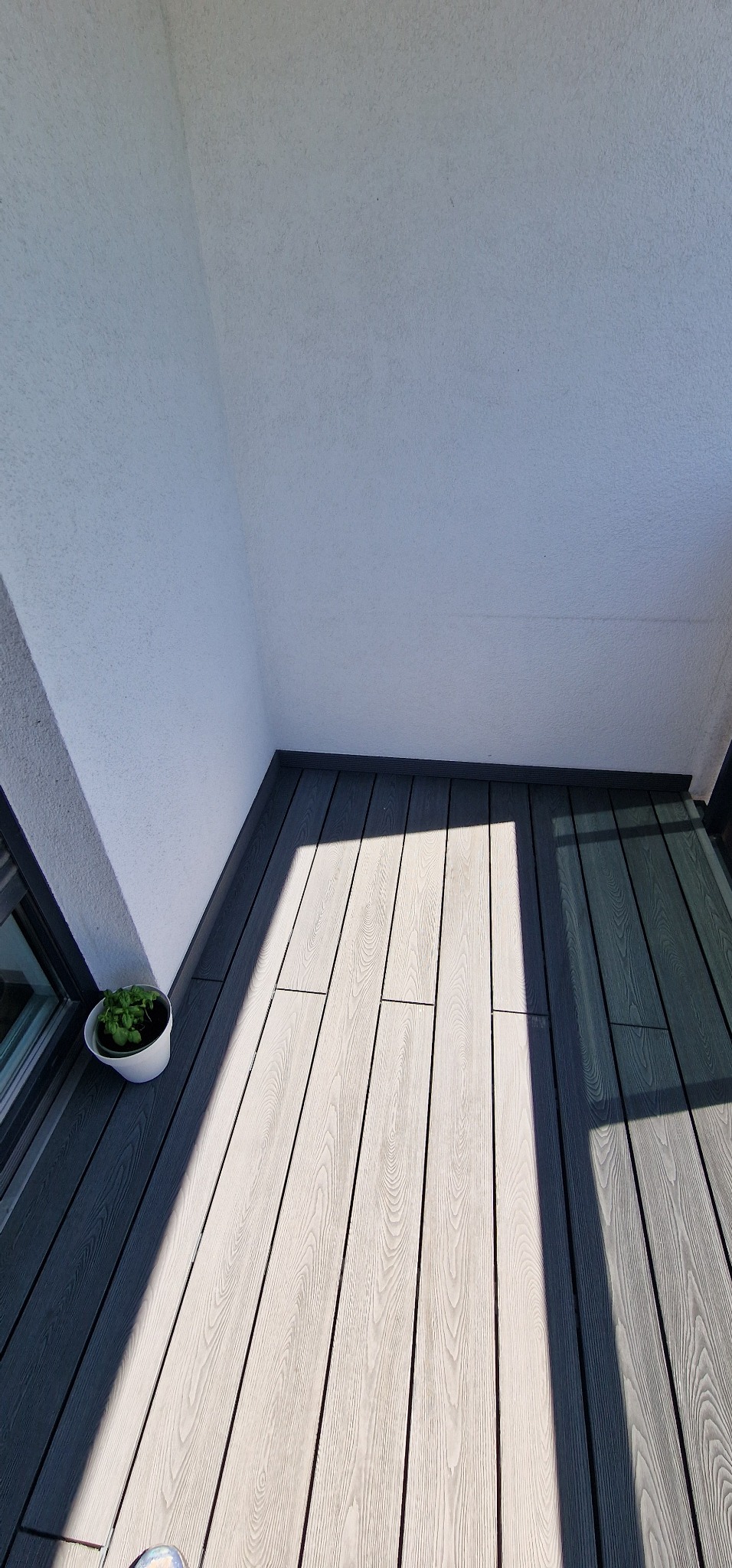 Nowoczesny balkon z szarym, kompozytowym deskowaniem. W lewym dolnym rogu doniczka z zieloną rośliną. Widoczne cienie tworzą geometryczne wzory na podłodze.