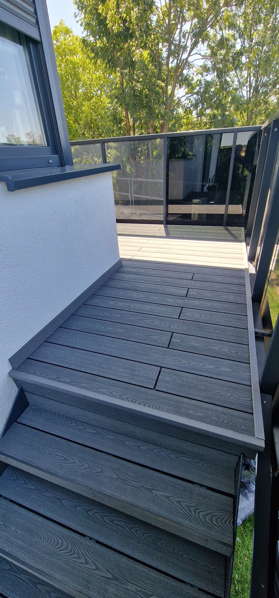 Nowoczesny taras z szarych desek kompozytowych, z niskimi schodami i szklaną balustradą, otoczony zielenią. Widok z góry, jasne oświetlenie.