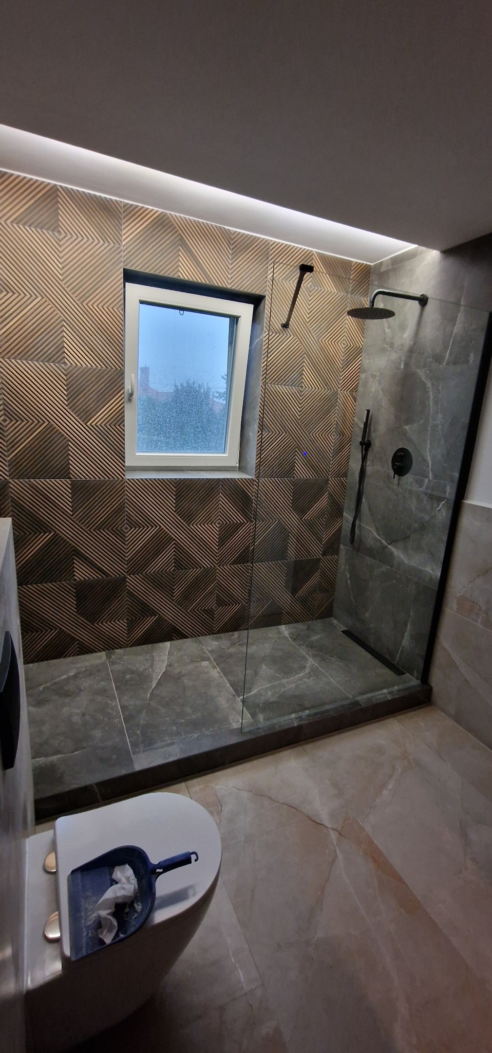 Nowoczesna łazienka z kabiną typu walk-in, wykończona płytkami w geometryczny wzór i imitacją marmuru, z oknem i podświetleniem LED. Widoczna toaleta z szufelką.