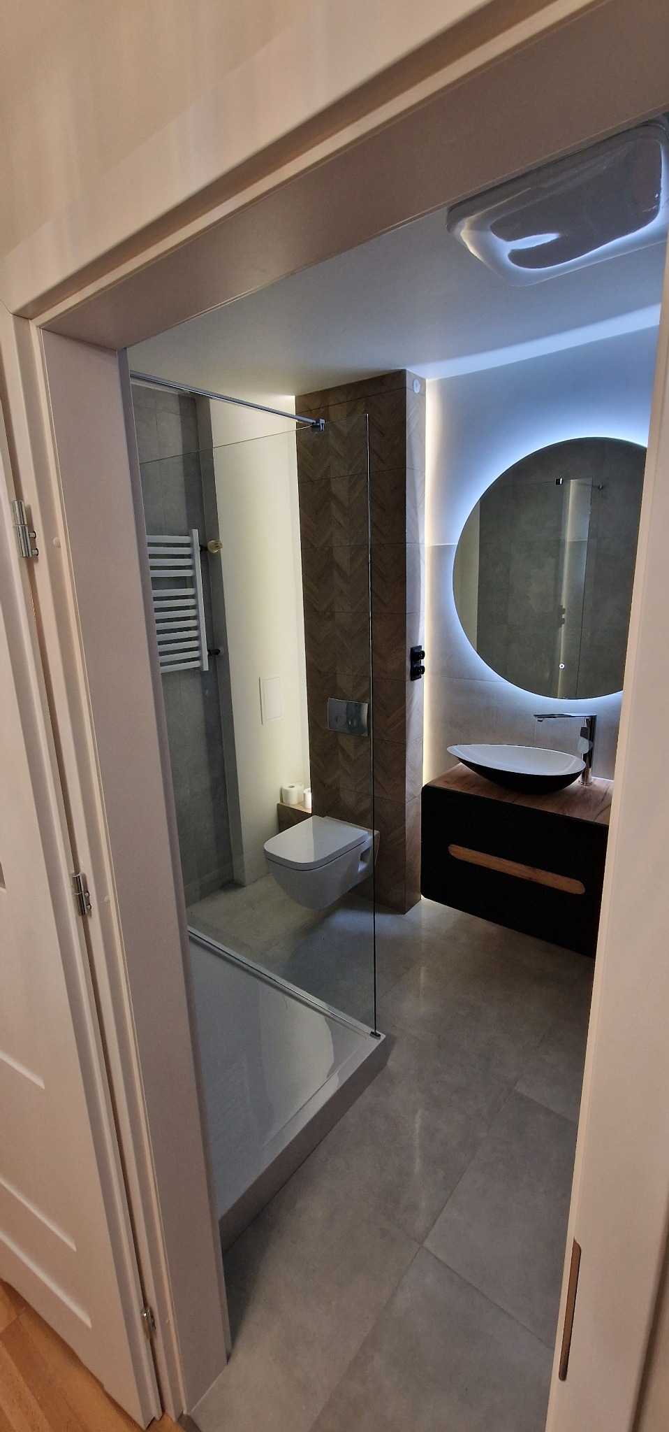 Nowoczesna łazienka po remoncie: kabina prysznicowa ze szklaną ścianą, podwieszana toaleta, umywalka na szafce z okrągłym lustrem podświetlanym LED.