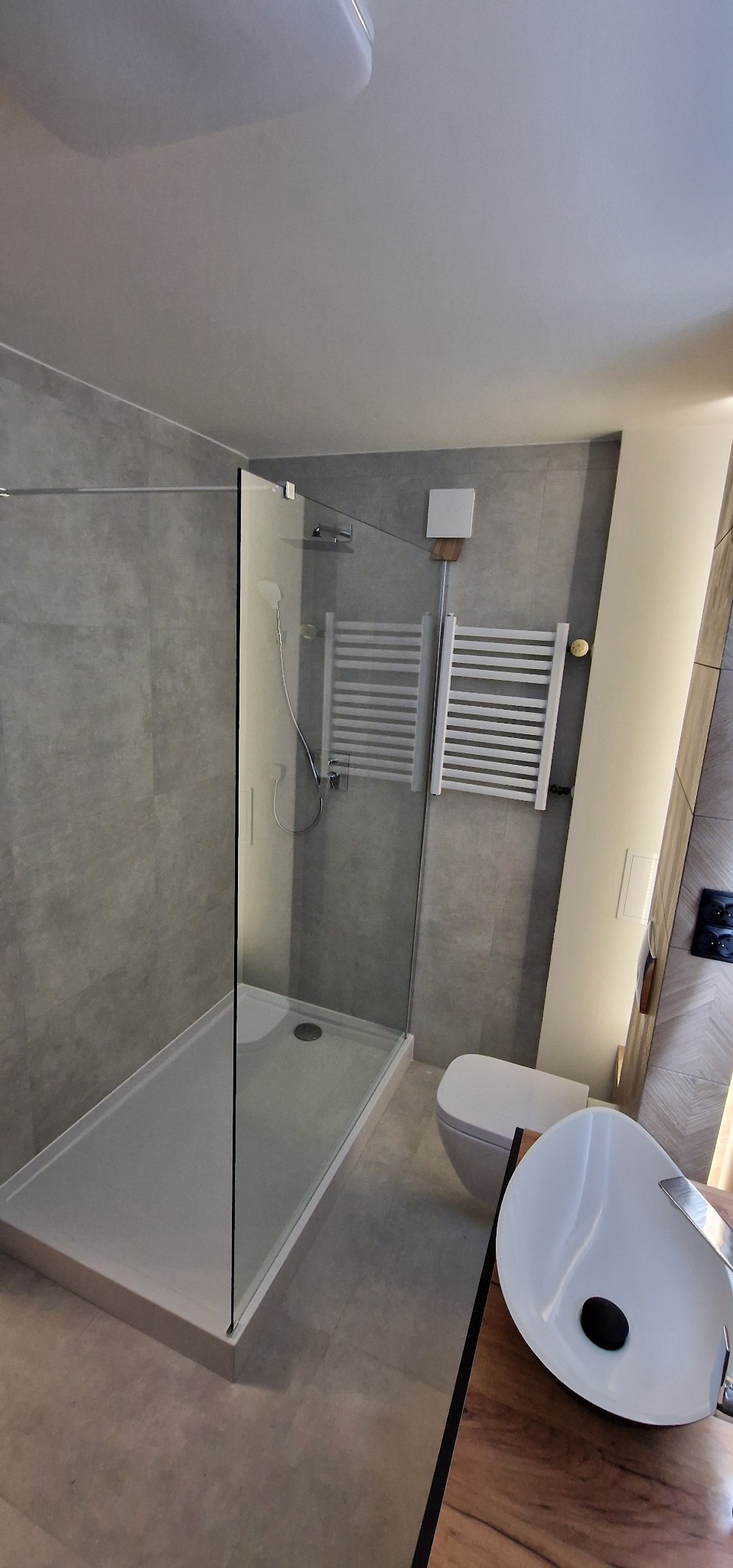 Nowoczesna łazienka w szarościach z kabiną prysznicową typu walk-in, grzejnikiem drabinkowym i umywalką nablatową na drewnianym blacie.