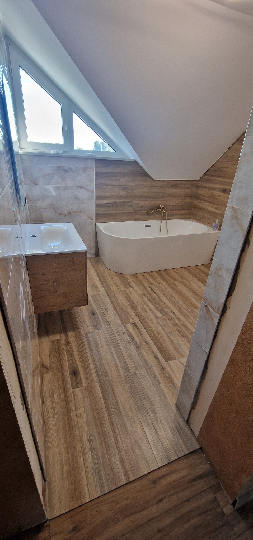 Jasna łazienka na poddaszu z wanną, umywalką z drewnianą szafką i oknem dachowym. Płytki imitujące drewno na podłodze i ścianach.