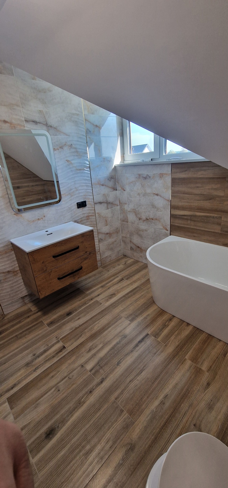 Nowoczesna łazienka na poddaszu z drewnopodobnymi płytkami, białą wanną i szafką z umywalką. Uwagę przyciąga lustro z podświetleniem LED.