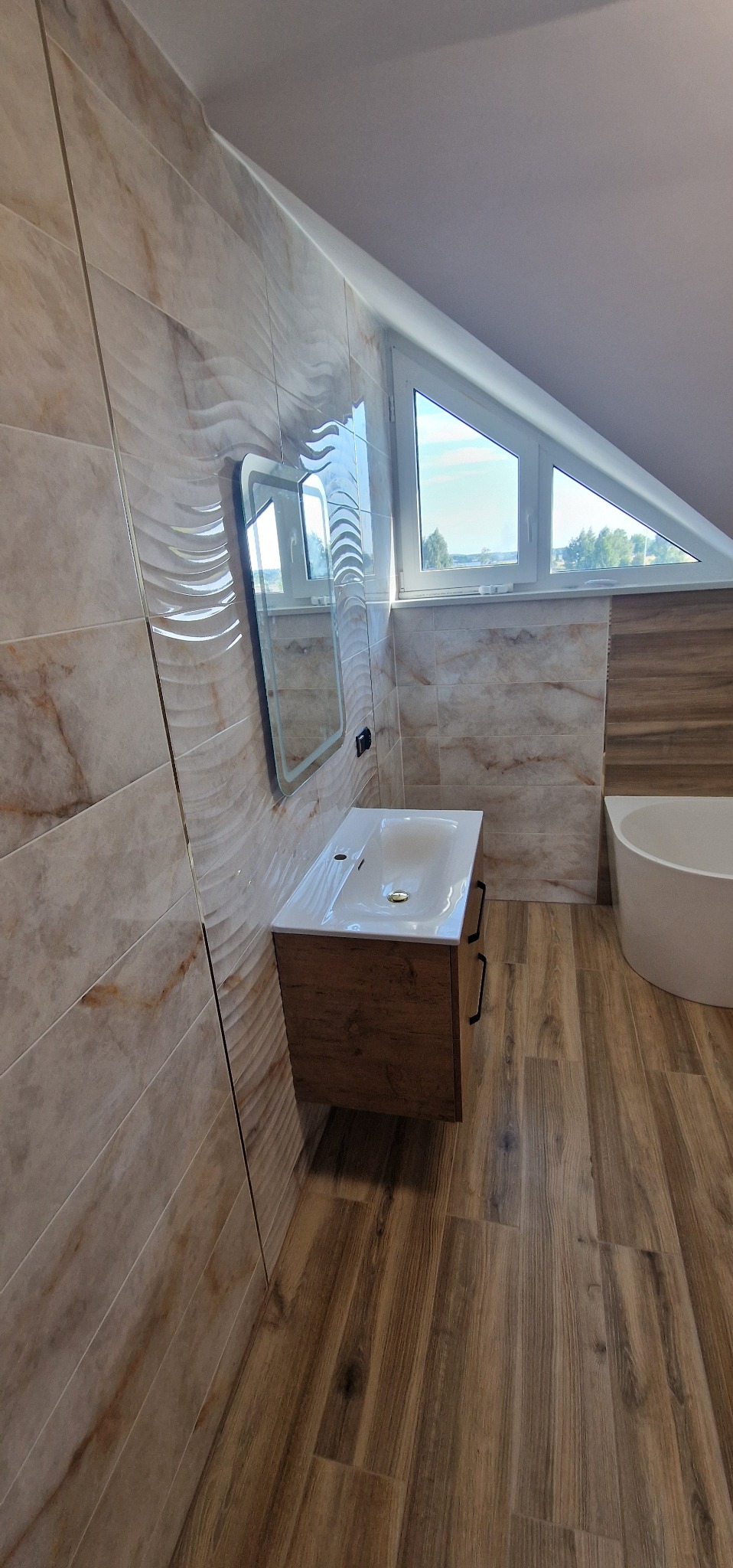Wykończona łazienka na poddaszu z drewnopodobną podłogą i beżowymi płytkami. Umywalka z szafką w kolorze drewna i lustro z podświetleniem. Okno dachowe trójkątne.