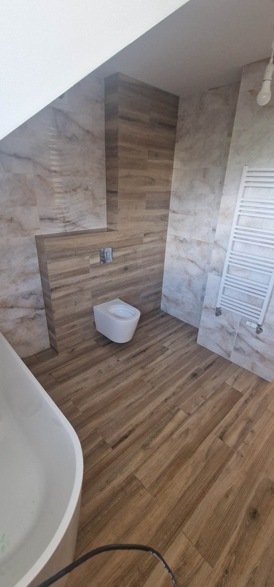 Nowoczesna łazienka po remoncie: drewnopodobne płytki na podłodze i ścianach, wisząca toaleta, biały grzejnik drabinkowy. Ukośny sufit.