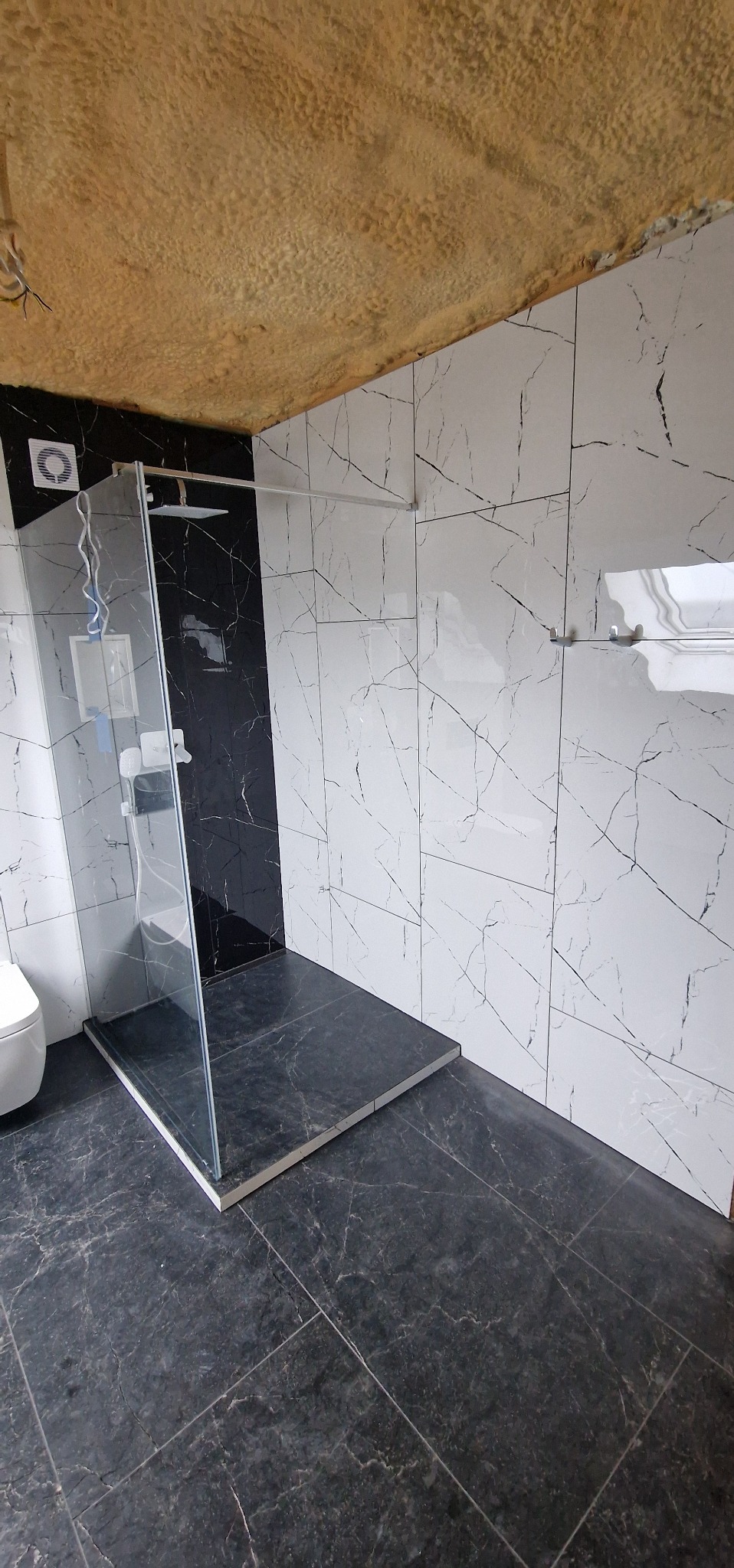 Wykończona łazienka z kabiną typu walk-in, czarną podłogą i białymi płytkami ściennymi z czarnymi żyłkami. Brodzik z płytek z białym rantem. Widoczna wentylacja.