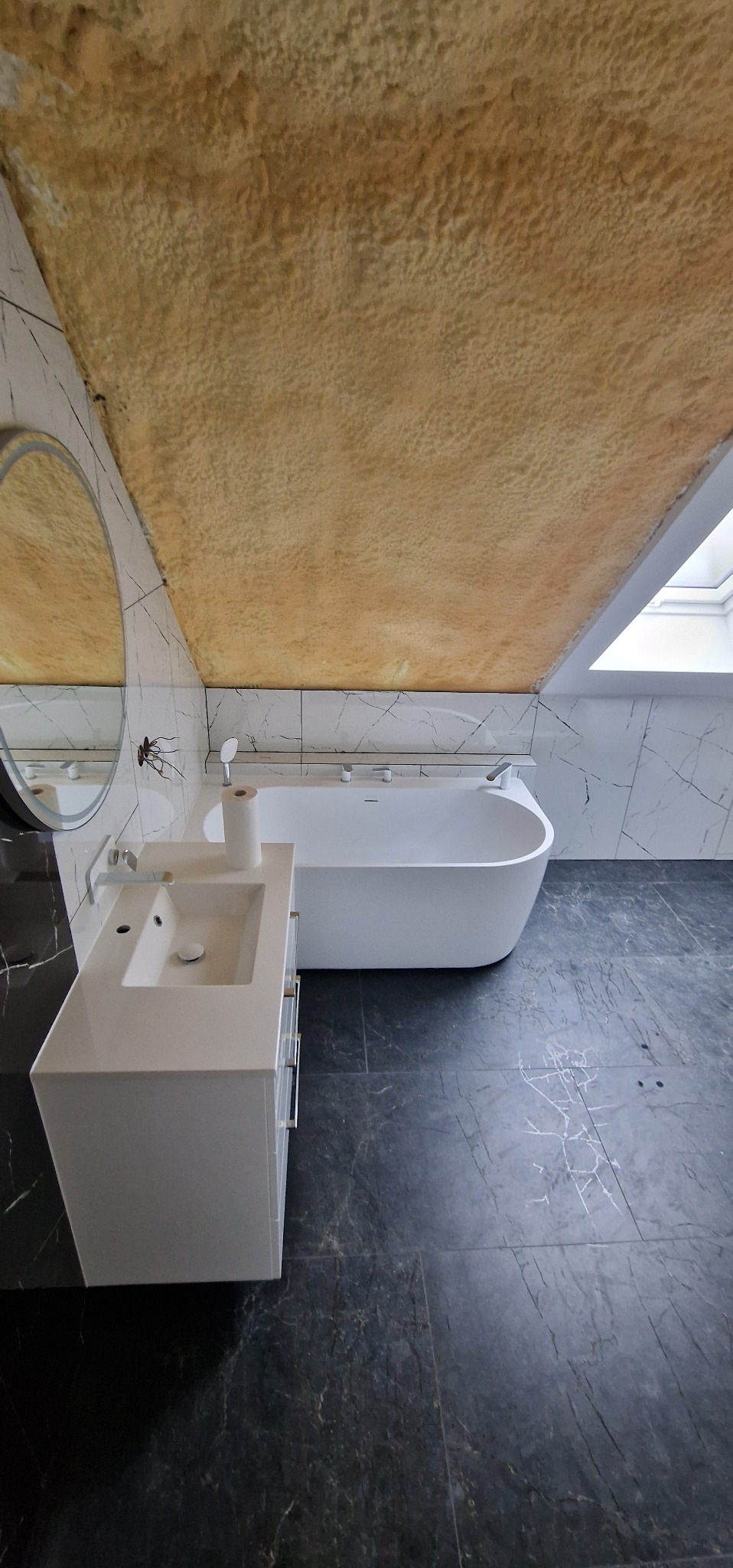 Nowoczesna łazienka na poddaszu z wanną, umywalką i marmurowymi płytkami. Sufit wykończony w beżowej strukturze. Okrągłe lustro z podświetleniem.