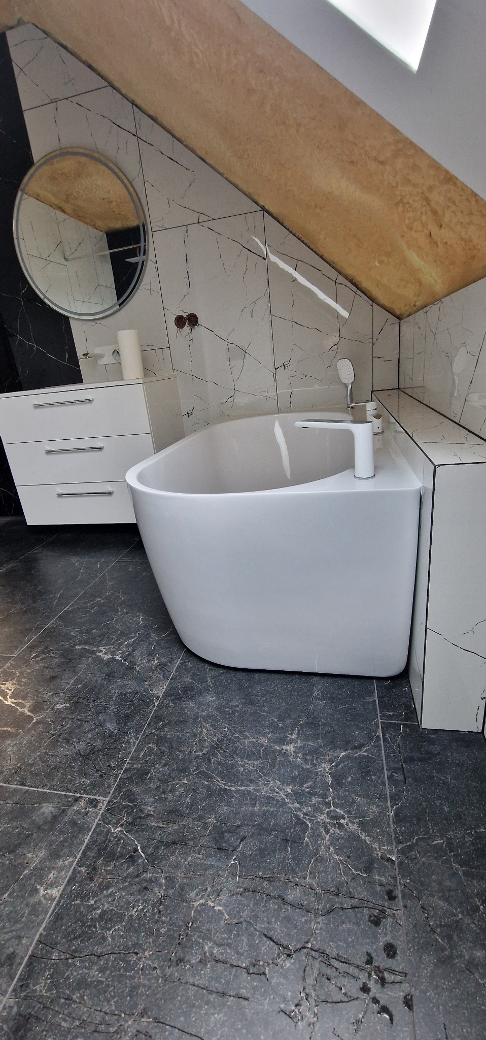 Nowoczesna łazienka na poddaszu: wolnostojąca wanna, marmurowe płytki, okrągłe lustro z podświetleniem i biała szafka z szufladami. Ukośny sufit z drewnianym wykończeniem.