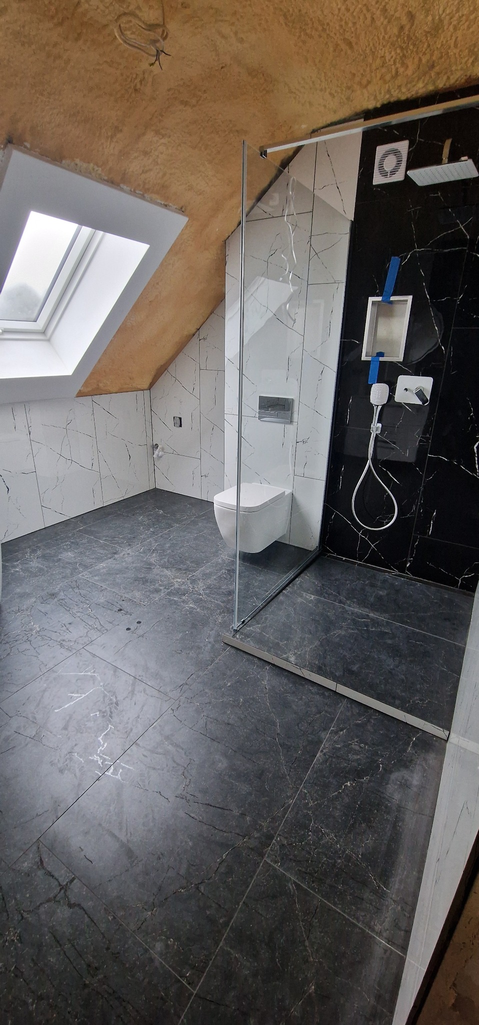 Wykończona łazienka na poddaszu z oknem dachowym. Czarne i białe płytki imitujące marmur, kabina prysznicowa ze szkła, podwieszana toaleta. Nowoczesny design.