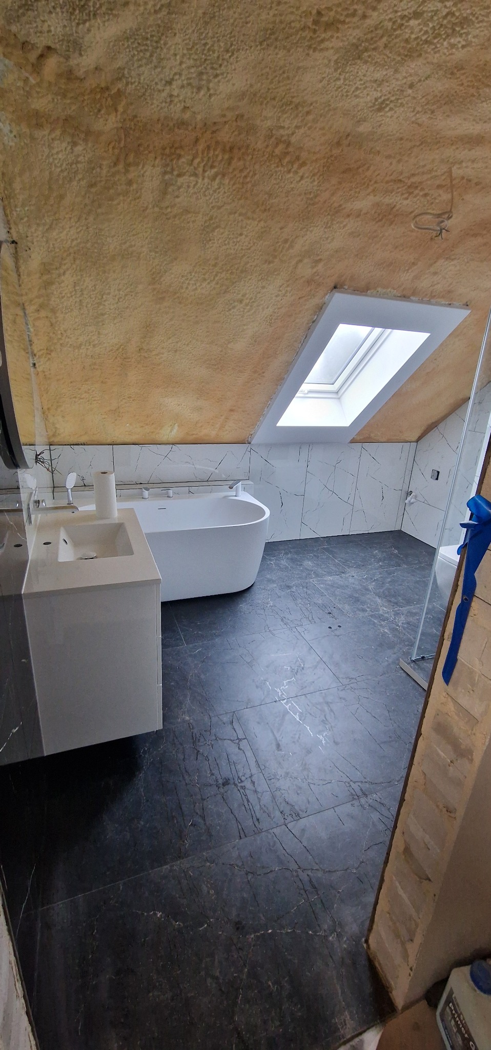 Wykończona łazienka na poddaszu z marmurową podłogą i ścianami, białą wanną i umywalką. Widoczne okno dachowe i skośny sufit z izolacją natryskową.