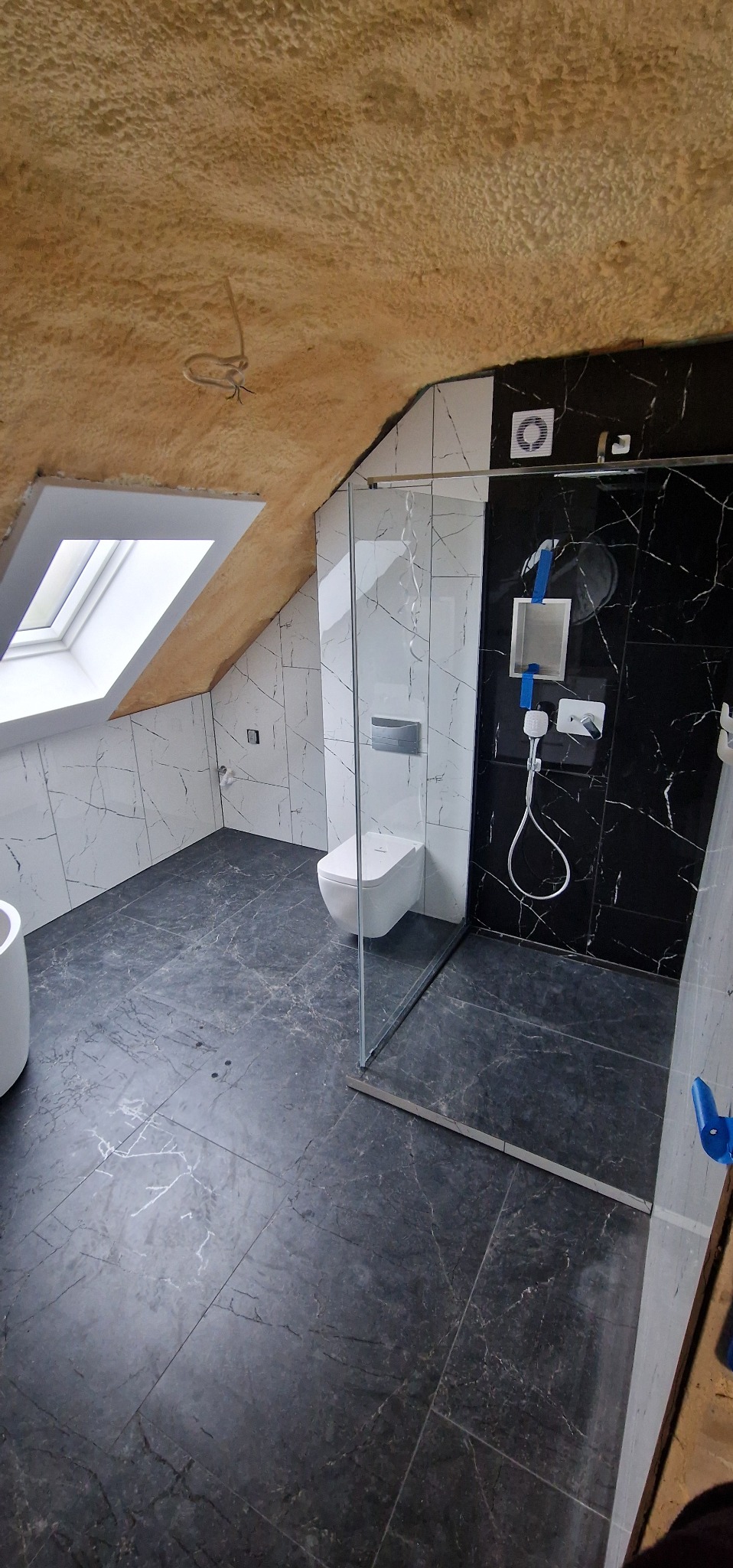 Wykończona łazienka na poddaszu z oknem dachowym. Czarne i białe marmurowe płytki, szklana kabina prysznicowa, wisząca toaleta. Sufit wykończony pianką.