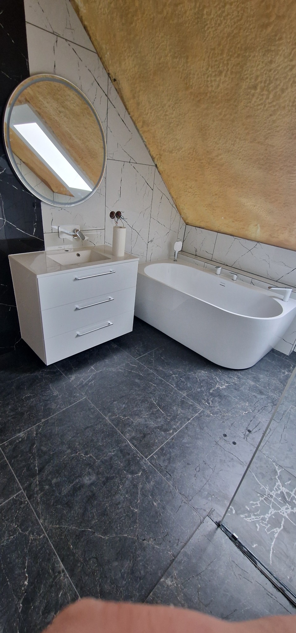 Nowoczesna łazienka na poddaszu: biała wanna wolnostojąca, szafka z umywalką i okrągłe lustro na tle marmurowych płytek i skośnego, złocistego sufitu.