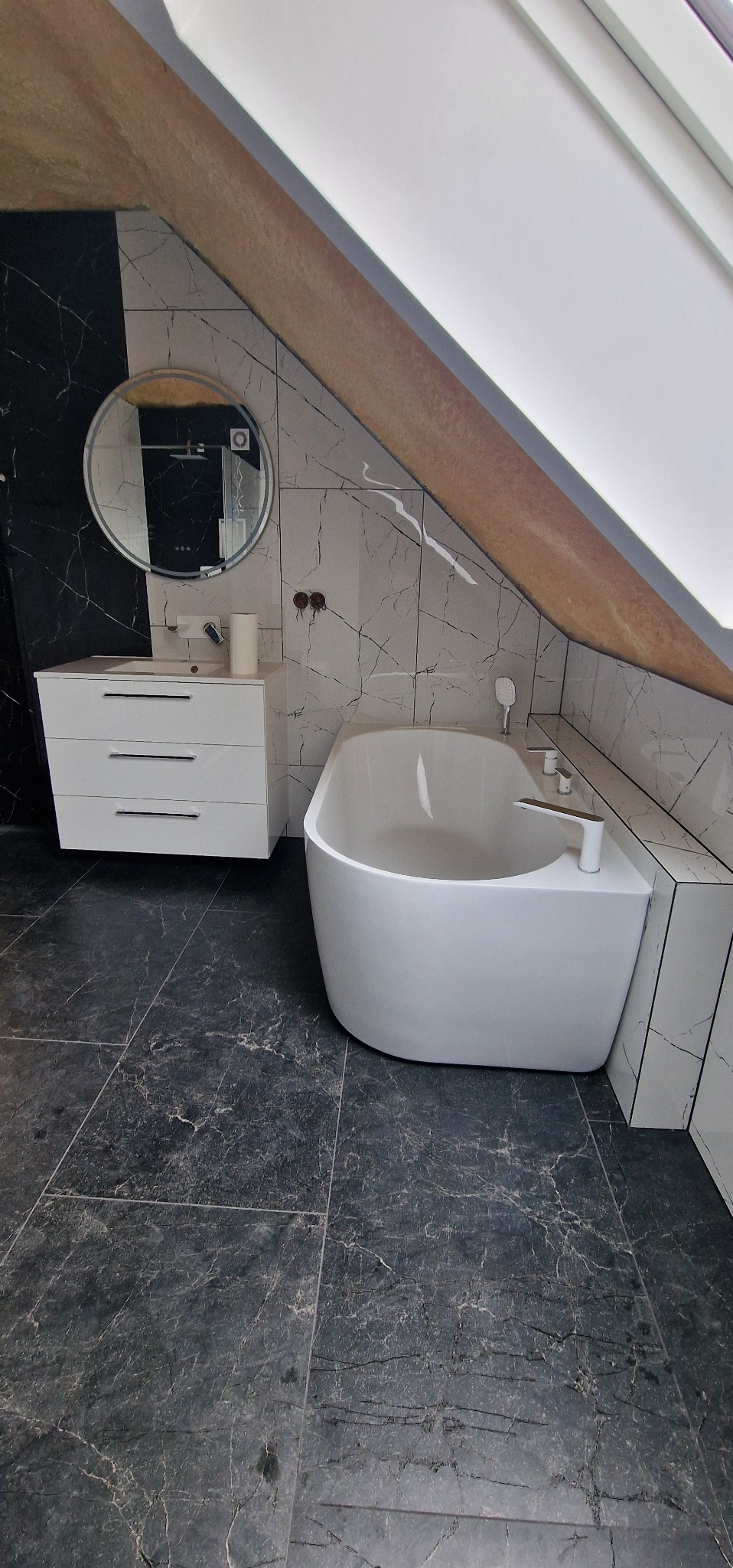 Nowoczesna łazienka na poddaszu z czarno-białymi marmurowymi płytkami, owalną wanną i białą szafką z umywalką. Okrągłe lustro i skośny sufit dodają unikalnego charakteru.