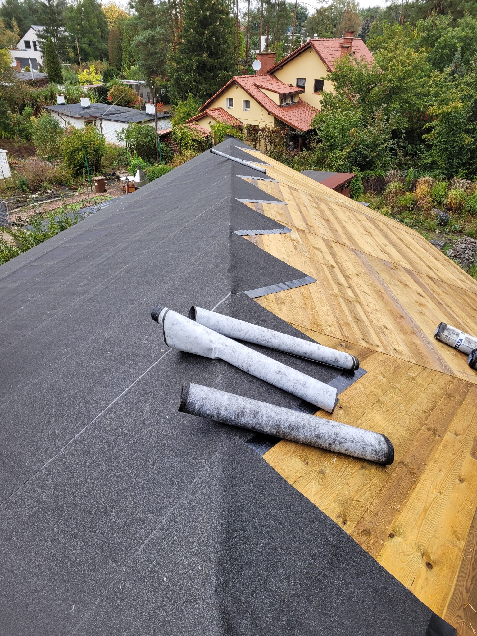Częściowo pokryty papą dach z widocznym deskowaniem i rolkami papy na tle zabudowy jednorodzinnej.
