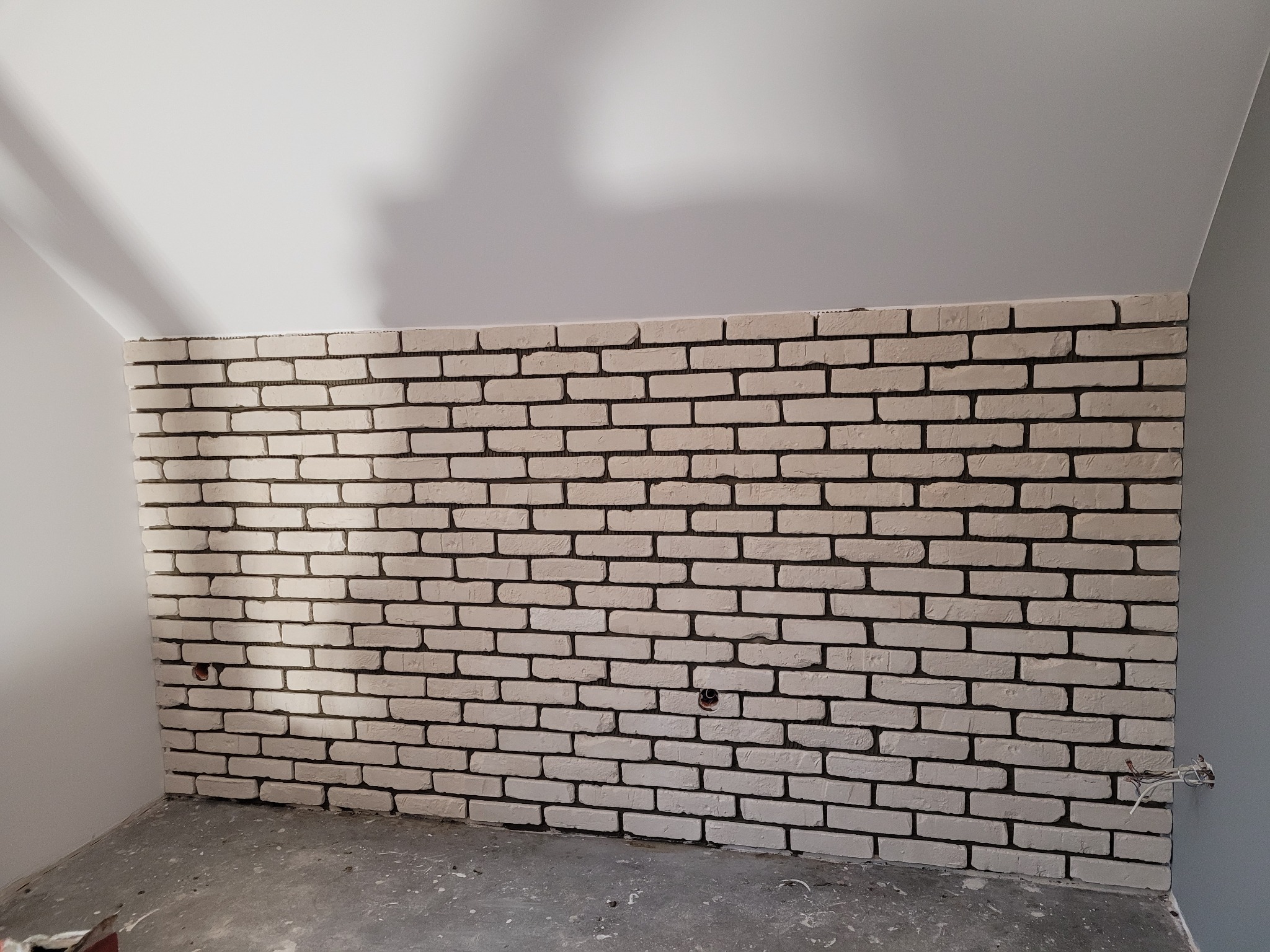 Ściana wykończona dekoracyjną cegłą w odcieniach bieli, widoczne otwory na gniazdka elektryczne, surowy beton na podłodze, ściana pod skosem