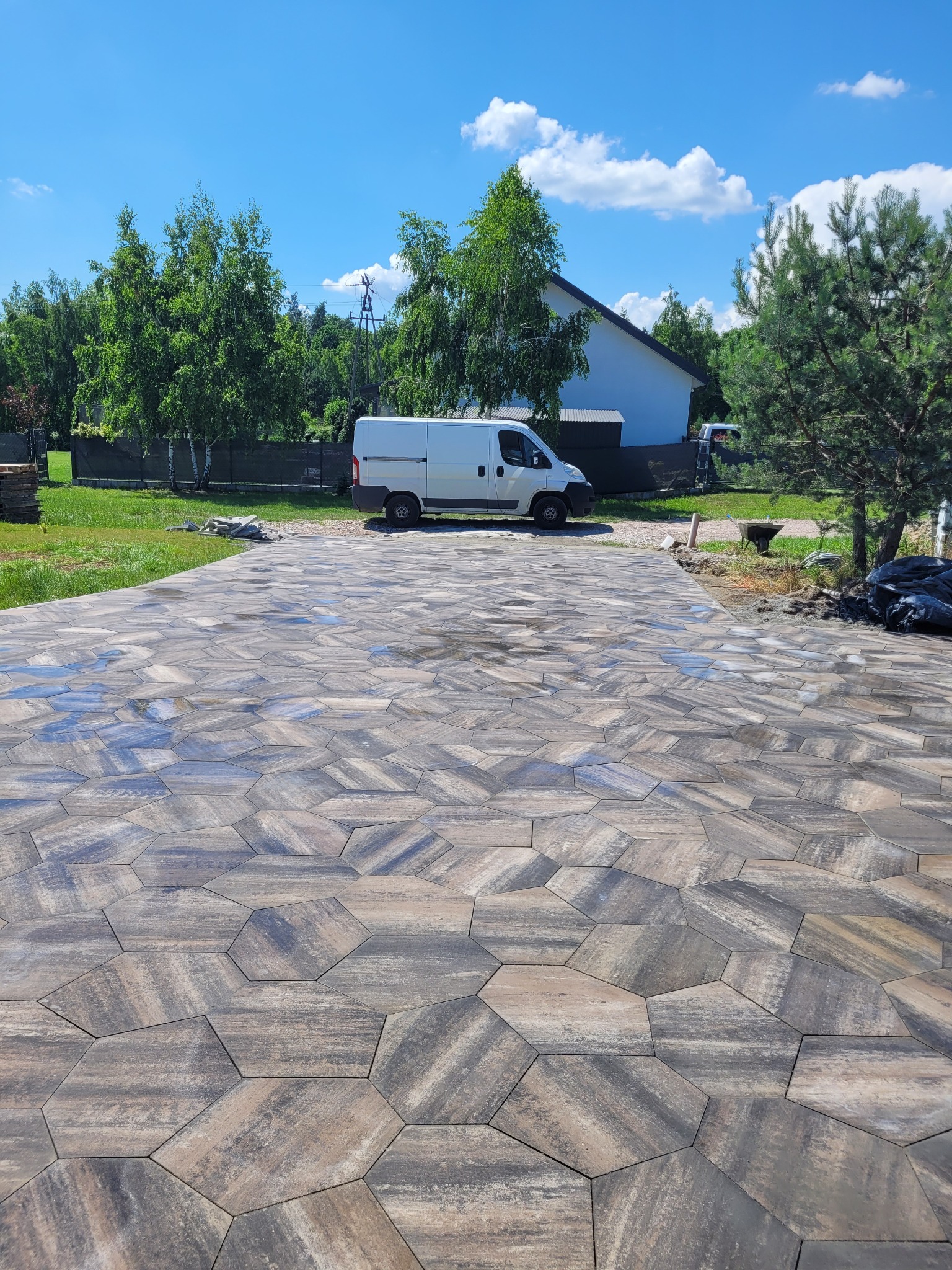 Ułożona nawierzchnia z heksagonalnych płyt w odcieniach szarości i beżu, z białym busem w tle i fragmentem domu, widok z niskiej perspektywy.