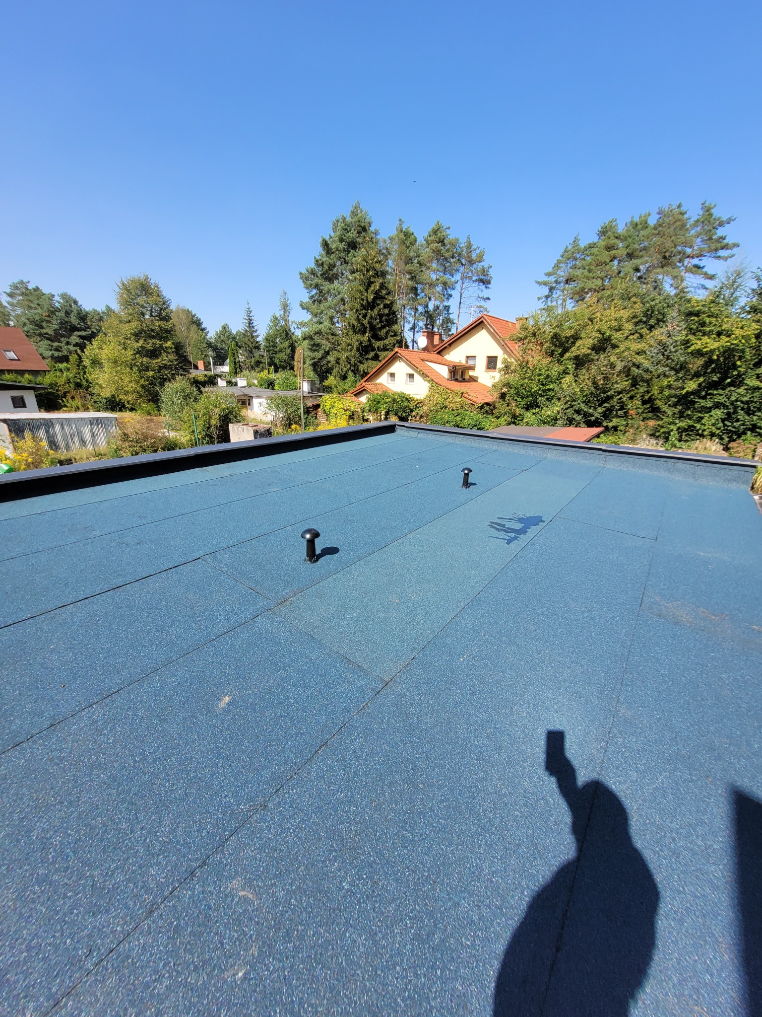 Niebieski dach płaski z papy termozgrzewalnej z dwoma kominkami wentylacyjnymi, widok na domy jednorodzinne i drzewa w tle, cień osoby robiącej zdjęcie.